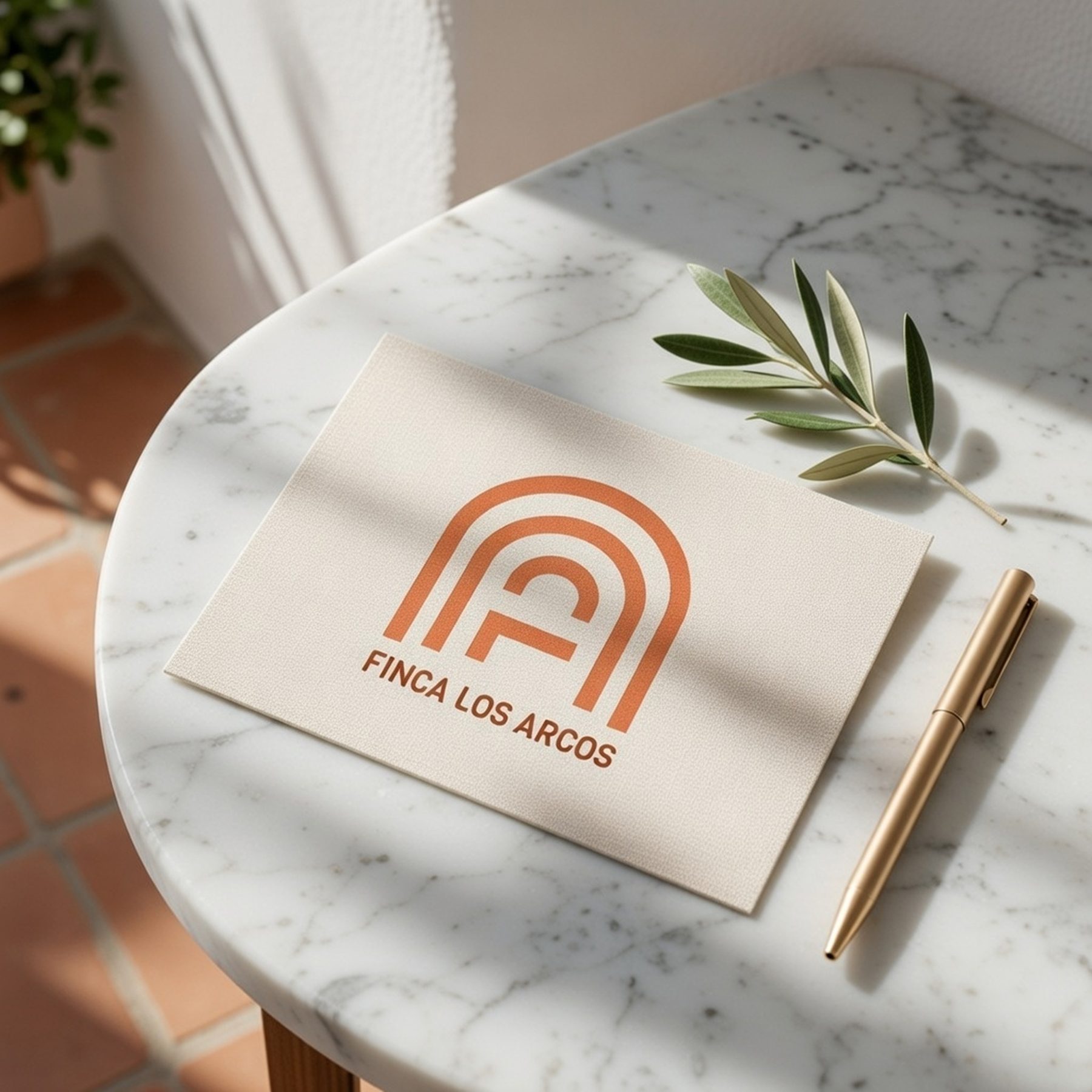 A cream textured card with the terracotta arched Finca Los Arcos logo on a white marble table, beside a gold pen and a fresh olive sprig in sunlight.