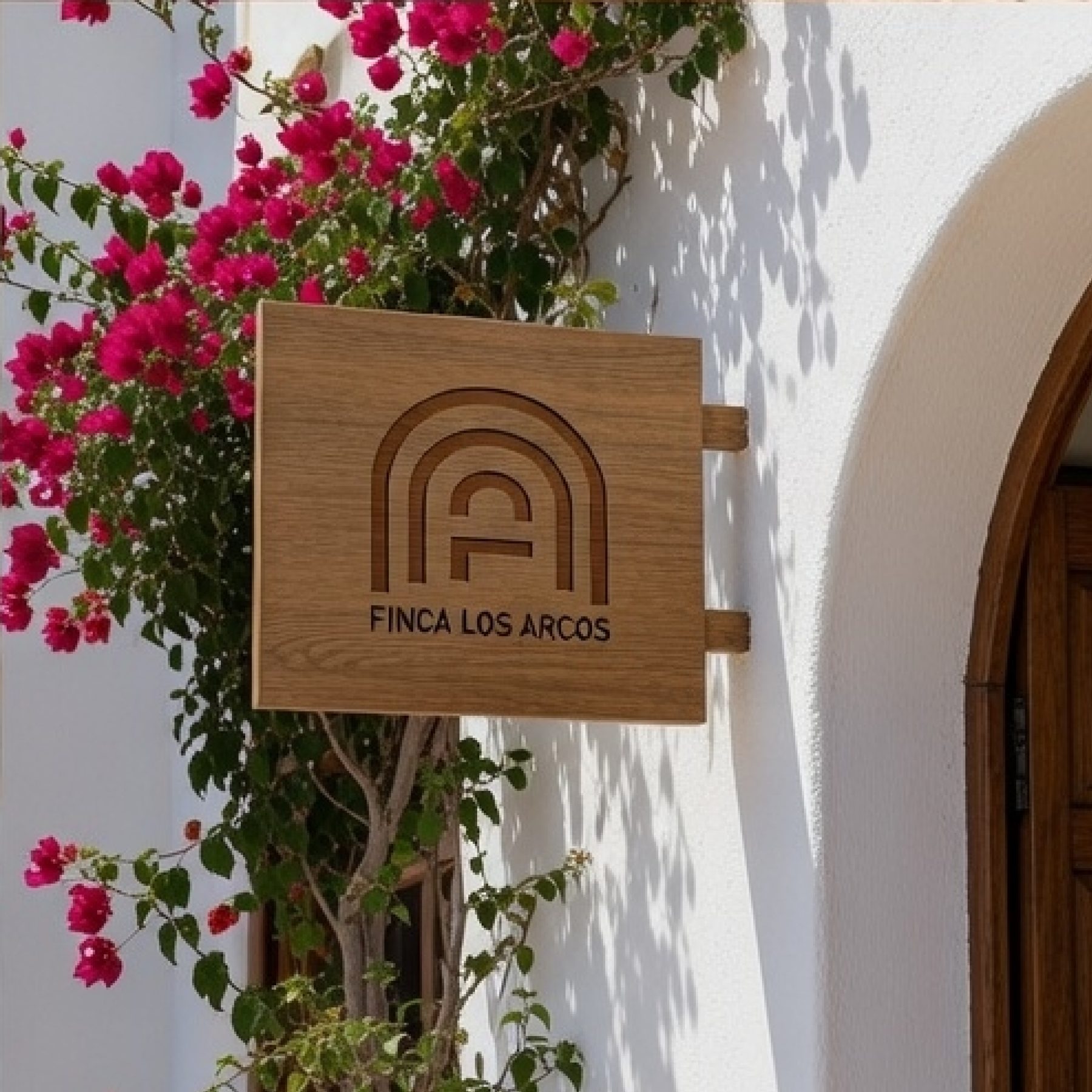 A square light-wood sign engraved with the Finca Los Arcos logo and name, mounted on a whitewashed wall next to pink bougainvillea and a dark wood arched doorway.